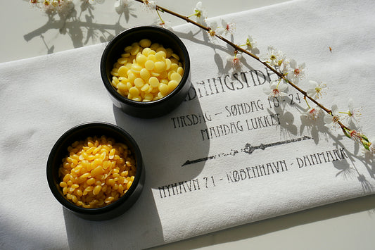 Zwei schwarze Schälchen auf einem weißen Geschirrtuch eines ist gefüllt mit Kakaobutterpellets, das andre mit goldenen Bienenwachspellets. Daneben liegt ein weiß blühender Zweig