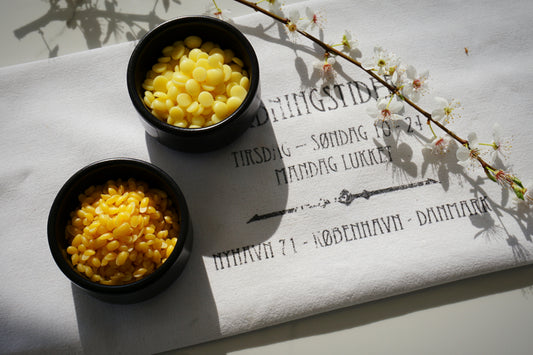 Zwei schwarze Schälchen auf einem weißen Geschirrtuch eines ist gefüllt mit Kakaobutterpellets, das andre mit goldenen Bienenwachspellets. Daneben liegt ein weiß blühender Zweig