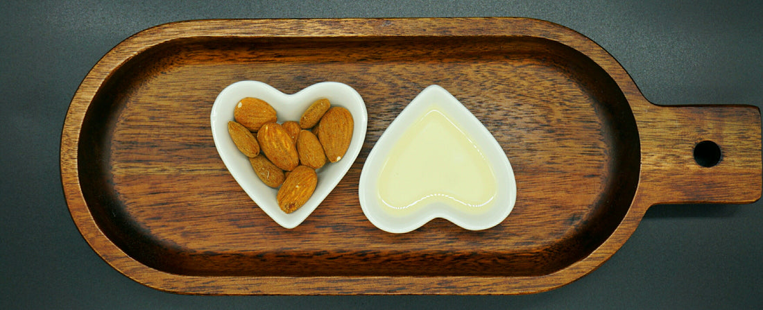 Zwei kleine weiße Glasschüsseln in Herzform, eine gefüllt mit Öl, die andere mit Mandeln auf einem ovalen Holzbrett